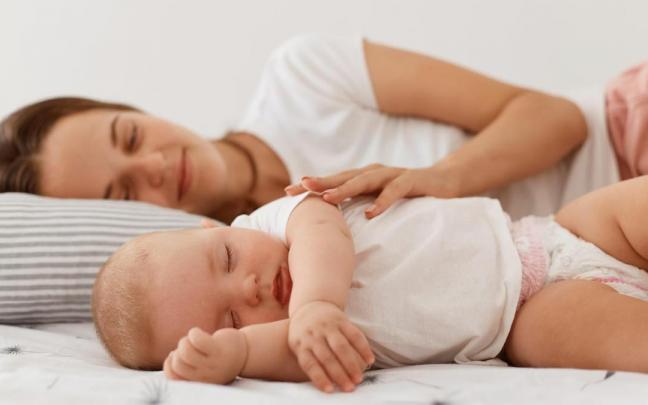 Una mujer acostada en la cama junto a su beb&eacute; dormido.