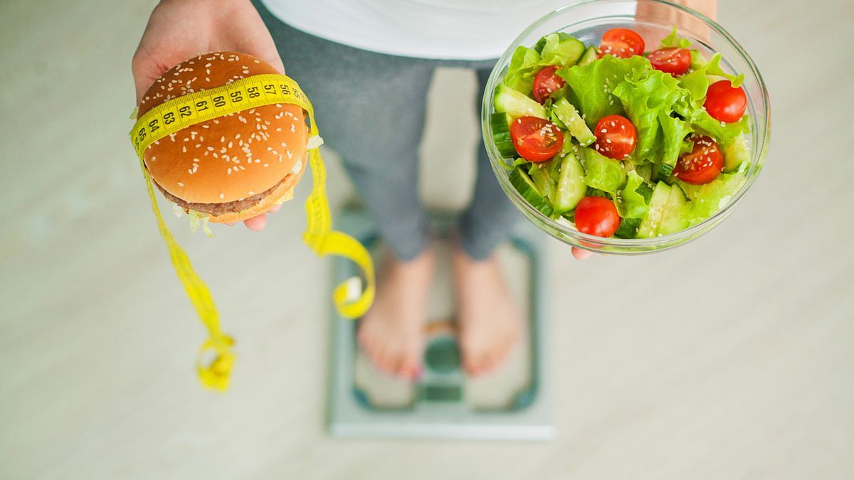 Una mujer subida a una báscula muestra en una mano una ensalada y en otra una hamburguesa.