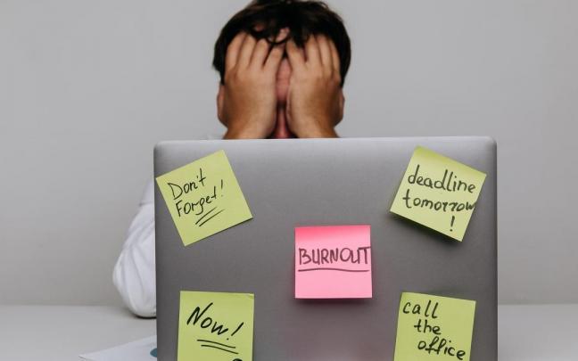 Un hombre agotado tras un portátil lleno de poss-it en uno de los cuales se lee 'burnout'. Un hombre agotado tras un portátil lleno de poss-it en uno de los cuales se lee 'burnout'.