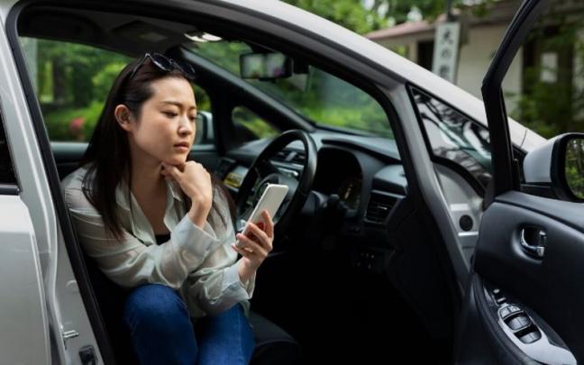 Mujer joven ojea su móvil sentada en el asiento delantero de un coche estacionado. Mujer joven ojea su móvil sentada en el asiento delantero de un coche estacionado.