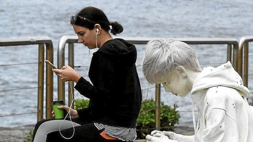 Una joven mira su teléfono móvil sentada junto a una escultura de un chico jugando con una videoconsola.