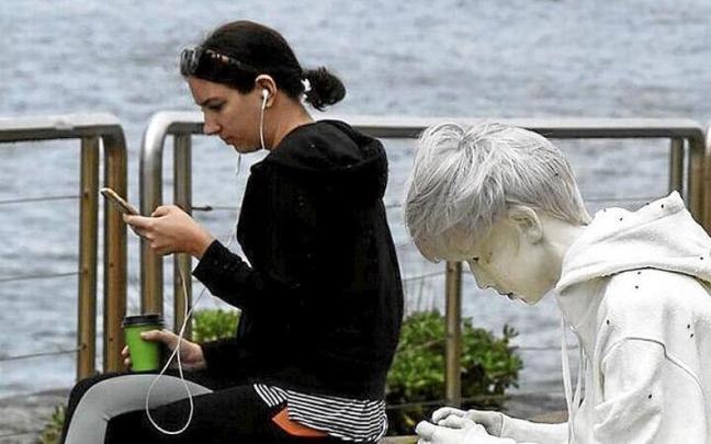 Una joven mira su teléfono móvil sentada junto a una escultura de un chico jugando con una videoconsola.
