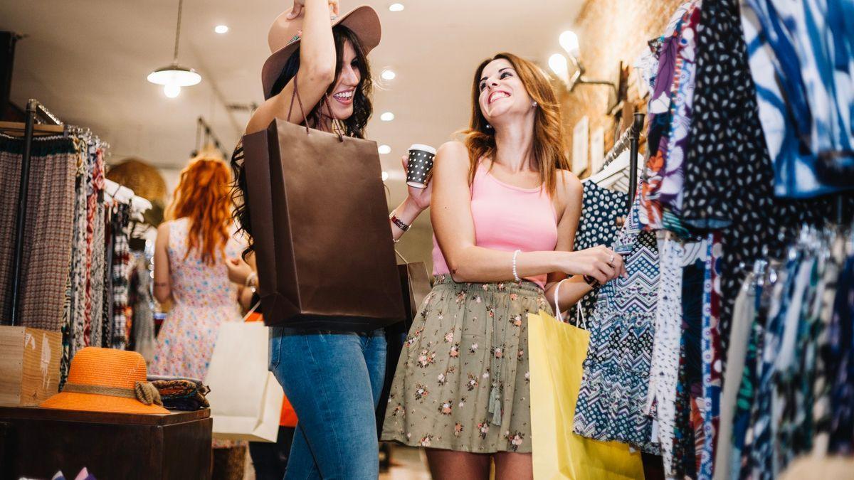 Dos mujeres jóvenes miran ropa en una tienda mientras beben café.