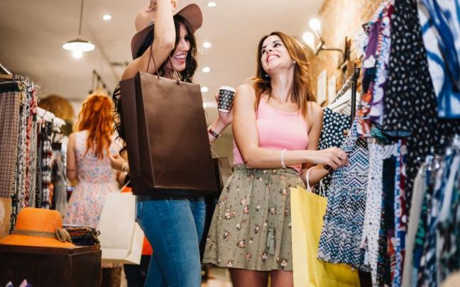 Dos mujeres j&oacute;venes miran ropa en una tienda mientras beben caf&eacute;.