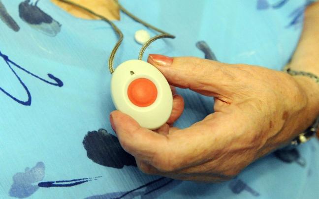 Una mujer muestra su medallón de teleasistencia con su característico botón rojo. Ahora también se distribuye como pulsera. Una mujer muestra su medallón de teleasistencia con su característico botón rojo. Ahora también se distribuye como pulsera.