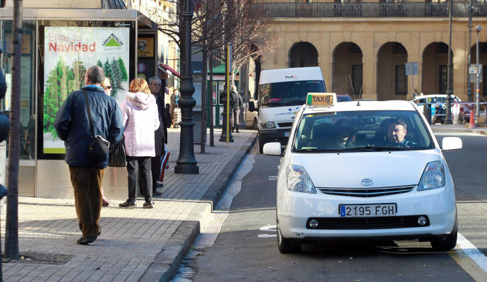 Usuarios esperando en una marquesina la llegada de una villavesa mientras un taxi se acerca