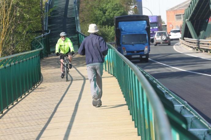 Un hombre y un ciclista pasean por la pasarela peatonal de Landaben que conecta el Parque Fluvial de Pamplona con el de la Comarca en Arazuri, junto a la carretera del polígono (PA-30).