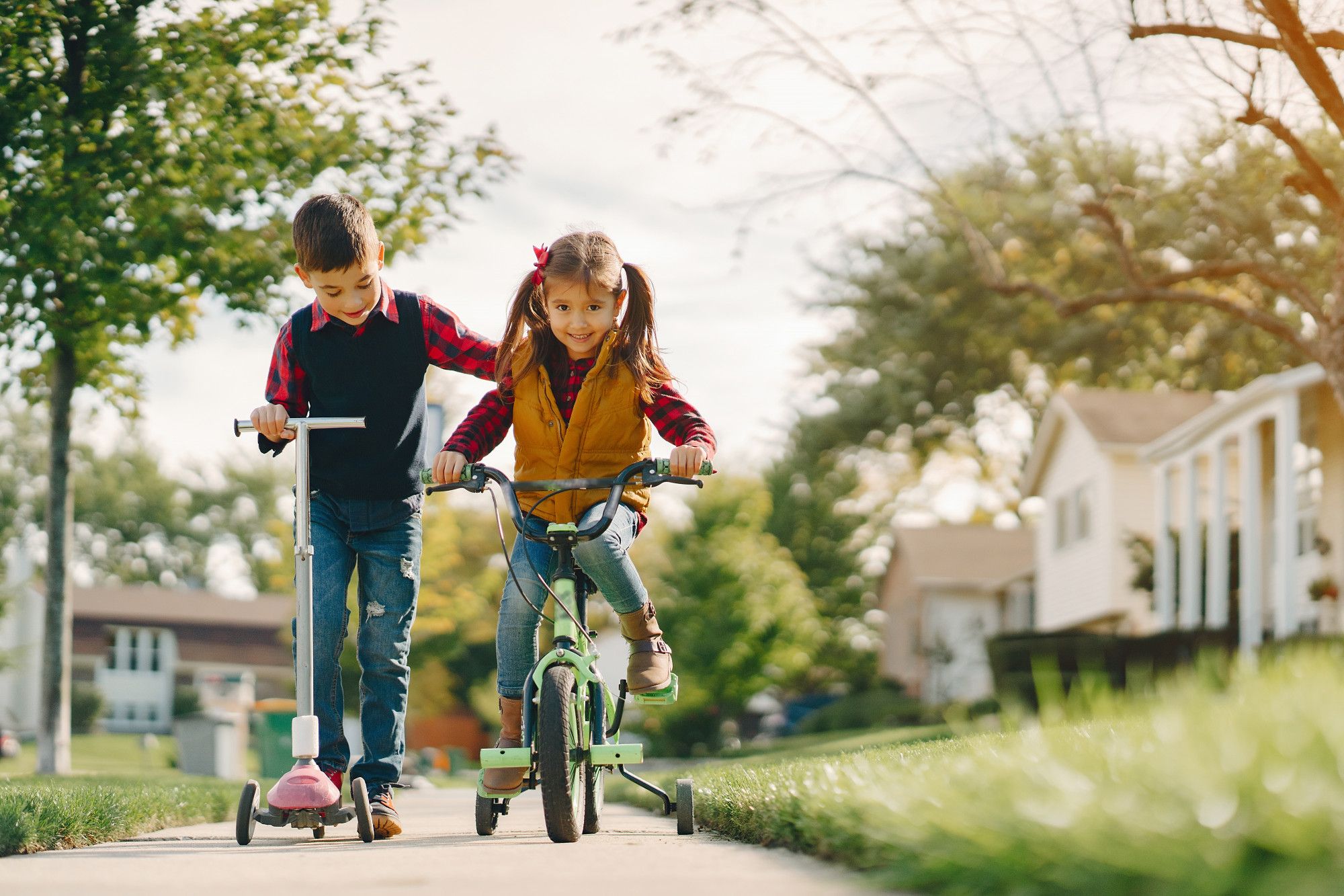 Un niño en patinete ayuda a una niña montada en una bici con ruedines.