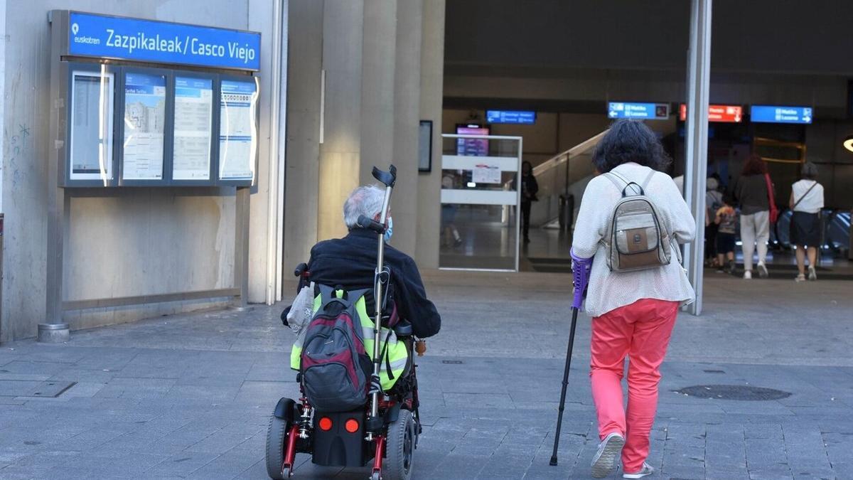 Una persona camina ayudada por una muleta junto a otra, en silla de ruedas, en Bilbao.