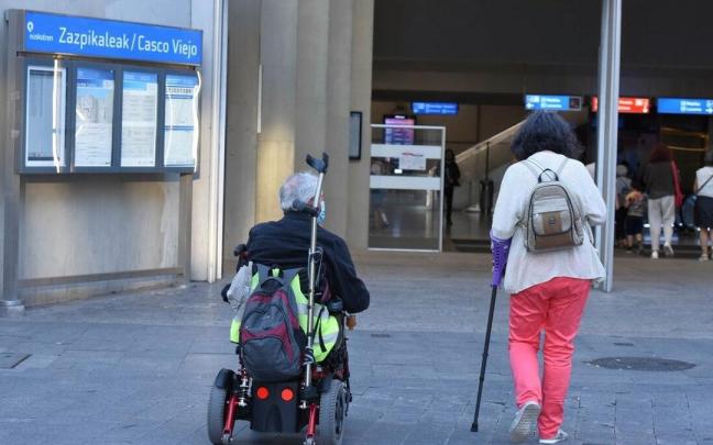 Una persona camina ayudada por una muleta junto a otra, en silla de ruedas, en Bilbao.