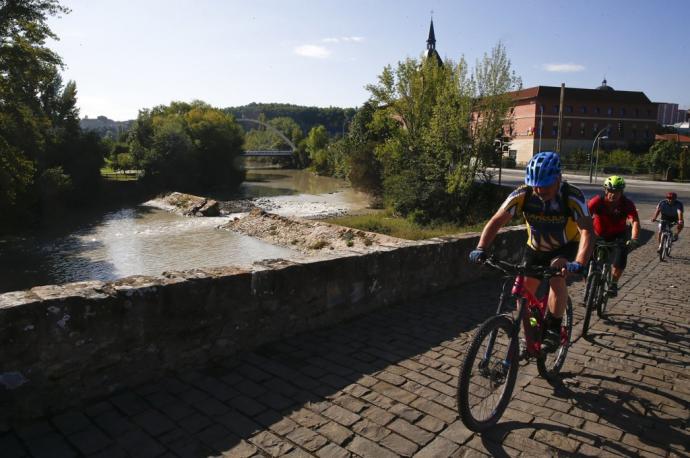 Ciclistas cruzando el puente de Santa Engracia. Detrás, la presa, rota y, al fondo, el puente de Oblatas.